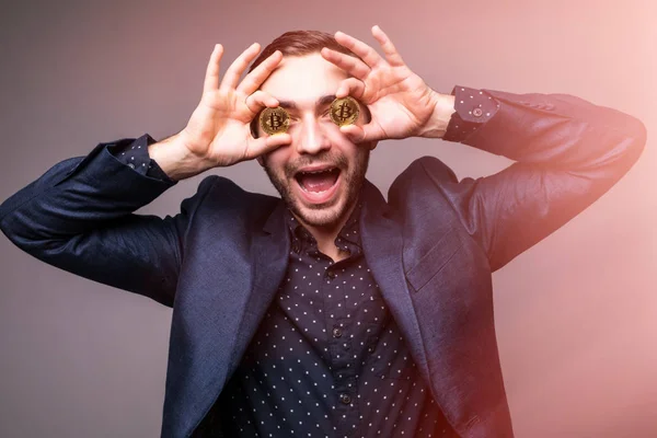Entusiasmado homem bonito de terno com bitcoins cobrir seus olhos isolados no fundo escuro com luz respingo — Fotografia de Stock
