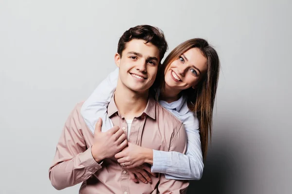 Портрет веселой молодой пары, обнимающейся стоя и глядя на камеру над серым — стоковое фото