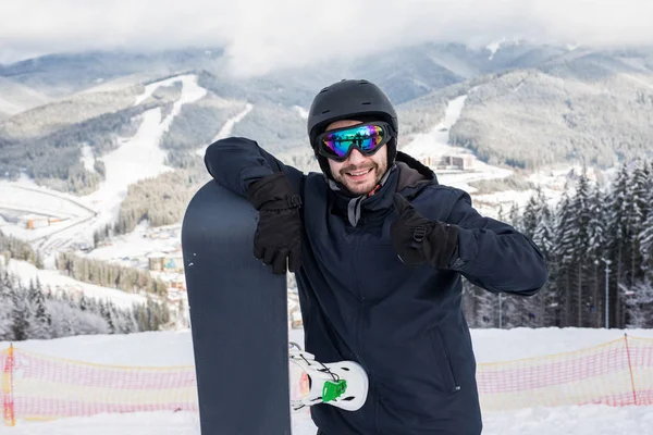 Hombre snowboarder de pie en la parte superior de la ladera nevada con snowboard, sonriendo a la cámara, mostrando los pulgares hacia arriba en la estación de esquí de invierno. Concepto de esquí y snowboard —  Fotos de Stock