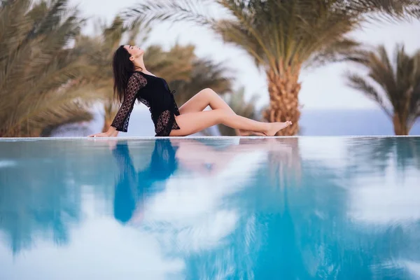 Hermosa chica sexy tumbada junto a la piscina infinita en la playa bajo las palmas —  Fotos de Stock