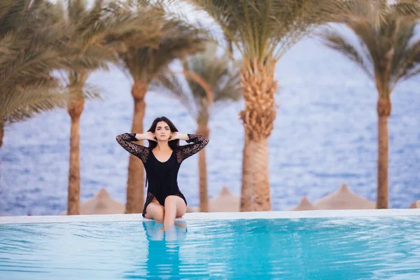 Una mujer joven está sentada junto al borde de una piscina infinita junto al océano y se relaja sobre el fondo de las palmeras —  Fotos de Stock
