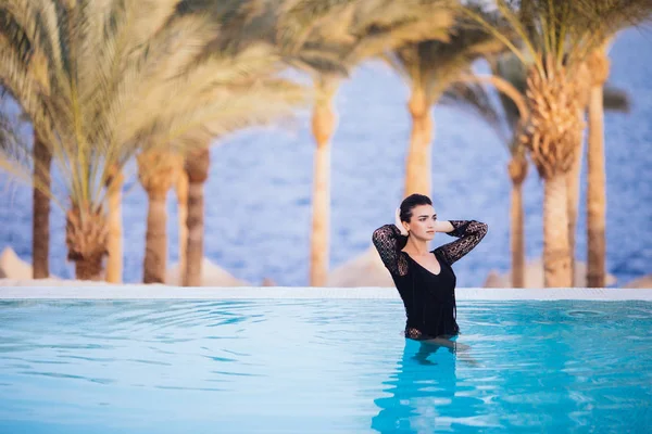 Mujer relajante en la piscina infinita posando en la cámara sobre el fondo de las palmas —  Fotos de Stock