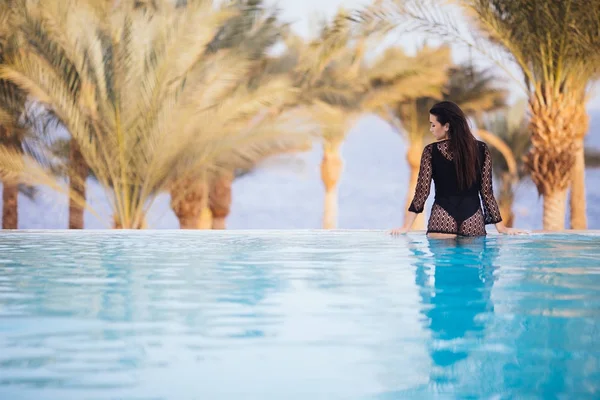 Parte posterior de la mujer sentada en el borde de la piscina infinita mirando a la vista del mar y las palmas. Vista trasera . —  Fotos de Stock