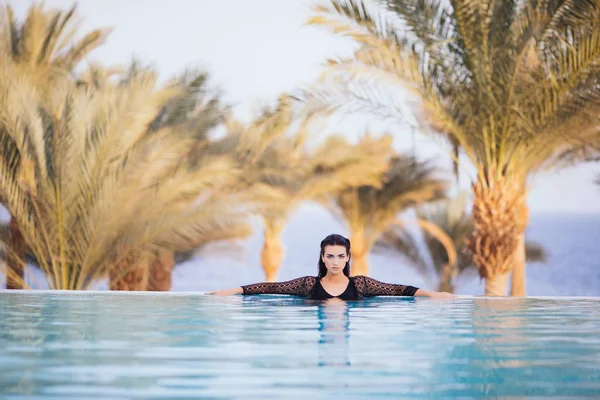 Retrato morena mujer relajarse en el agua de la piscina infinitocon las manos en el borde complejo de lujo, infinito vista al mar —  Fotos de Stock