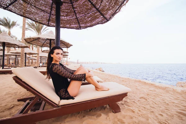 O femeie tânără frumoasă, cu picioare sexy și corp, care se bronzează la soare pe un scaun de punte pe plaja mării. Vocația de vară . — Fotografie, imagine de stoc