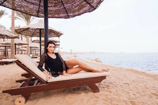 Jeune belle femme brune mince et sexy allongée sur une chaise longue sur la plage de la mer sous parapluie — Photo