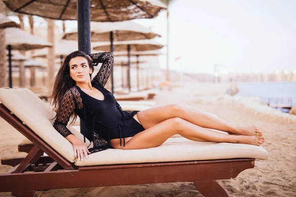 Jeune belle femme brune mince et sexy allongée sur une chaise longue sur la plage de la mer sous parapluie — Photo