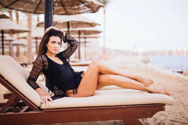 Joven hermosa mujer morena delgada y sexy tumbada en el sillón en la playa de mar bajo el paraguas —  Fotos de Stock