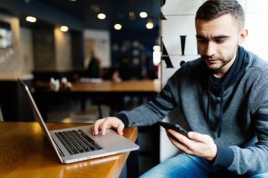 Mutlu genç gülümseyen ve kafede dizüstü bilgisayar ile oturup bir smartphone tutan adam. Portre adam kafede smartphone ile çıkıyor. Gülümseyen ve bir telefon mesajı yazma adam.