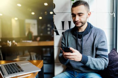 Seçici odak modern dijital smartphone aygıt tutarak ve sosyal ağlarda chat yapan adamın ellerinde. Bulanık genç adam kahve ve dizüstü bilgisayar ile masada otururken cep üzerinde SMS mesajı gönderme