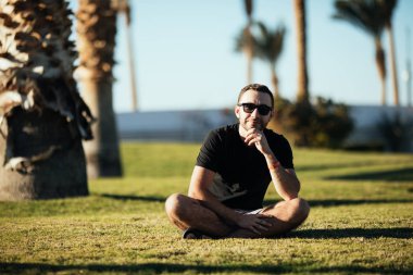 Yakışıklı genç adam bir kenara yaz meslek avucunda altında çimlerde oturan güneş gözlüğü bakan sakallı.