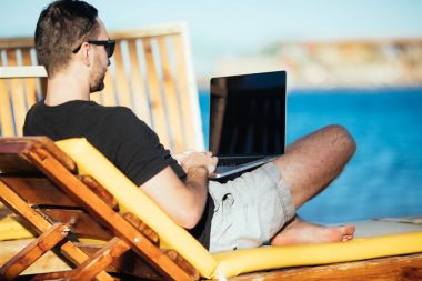 Yakışıklı adam sahilde onun güverte sandalyede otururken diz üstü siyah ekran ile yazarak