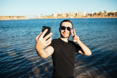 Deniz plaj arka plan üzerinde smartphone ile kendi portresi zafer işareti yapan genç yakışıklı erkek