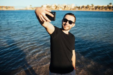 Mutlu genç adam tatilde plajda güneş gözlüğü deniz arka plan üzerinde selfie alarak gülüyor