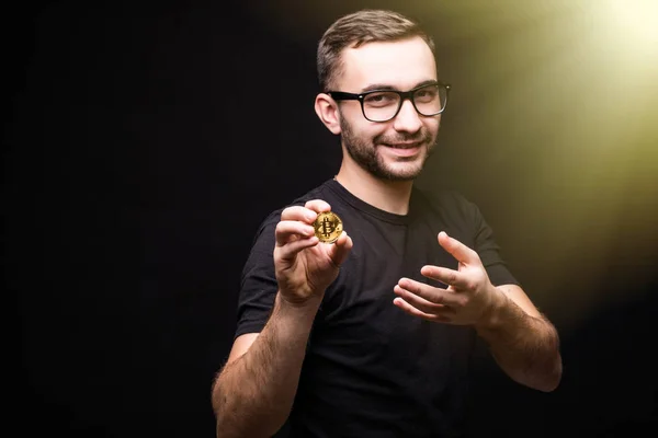 Hombre joven guapo en gafas de desgaste en camisa negra presente bitcoin aislado sobre fondo negro. Tonelada imagen . —  Fotos de Stock