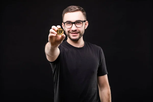 Portrét pohledný mladík v brýlích v černé košili ukázal zlatý bitcoin na kameře izolované na černém pozadí — Stock fotografie