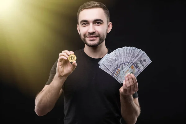 Junger gutaussehender Mann mit goldenem Bitcoin und Dollar-Bargeld in der anderen Hand isoliert auf schwarzem Hintergrund. Tonnenhaftes Image. — Stockfoto