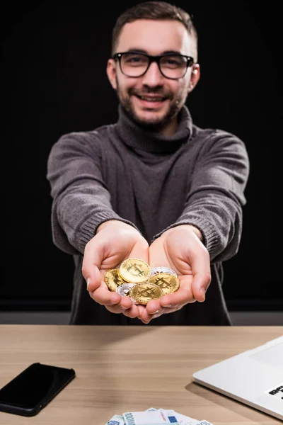 Portrét muže v brýlích ukazuje zlatá bitcoiny ve svých rukou na stůl izolované černém pozadí — Stock fotografie