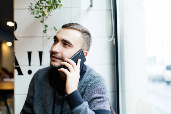 Genç işadamı, kafe ve kullanım smartphone masada oturan gri hırka giymiş sakallı. Holding smartphone dostum ve konuşmak. Adam Gadget'ı kullanma. — Stok fotoğraf
