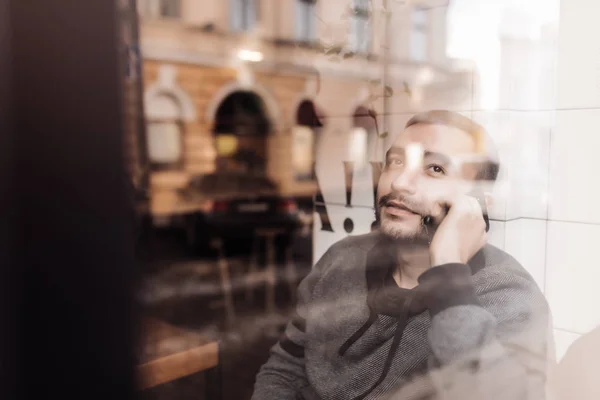 A gostar do telefonema dele. Bonito jovem falando no telefone celular e olhando através da janela com sorriso enquanto sentado no café — Fotografia de Stock