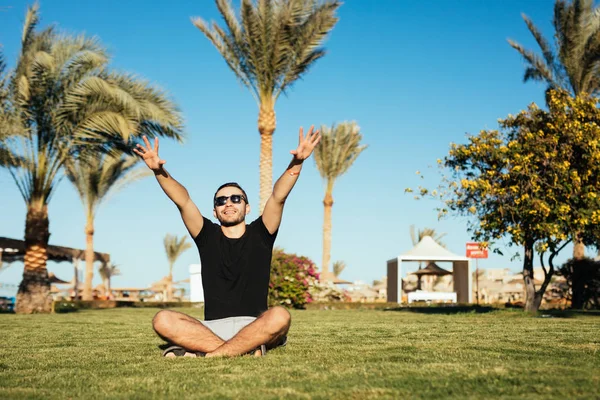 Красивый бородатый мужчина в солнечных очках сидит на зеленой траве и расслабиться насладиться летним призванием с поднятыми руками счастливой улыбкой на ладонях и синем фоне неба . — стоковое фото