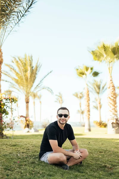 Красивый бородатый мужчина в солнечных очках сидит на траве под пальмами по летнему призванию . — стоковое фото