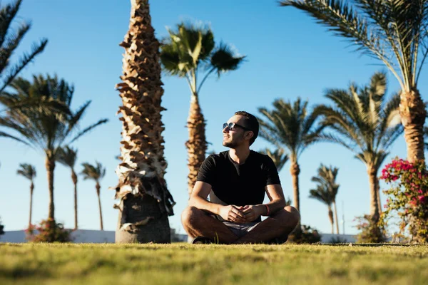 Красивый молодой бородатый мужчина в солнцезащитных очках сидит на траве под пальмами на роскошном курорте летнего отдыха — стоковое фото
