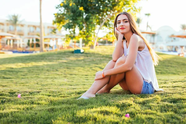 Hermosa joven sentada en la hierba en el parque bajo el sol —  Fotos de Stock