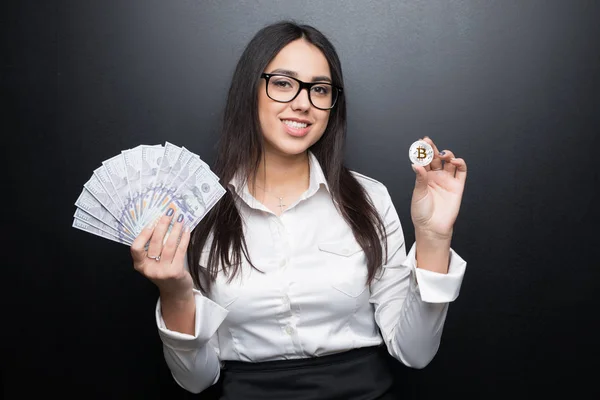 Νέων σύγχρονων επιτυχημένη γυναίκα των επιχειρήσεων σε ποτήρια κρατώντας ένα bitcoin και μετρητά χρήματα που απομονώνονται σε μαύρο φόντο — Φωτογραφία Αρχείου