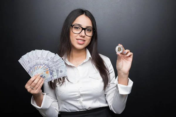 Молода сучасна успішна бізнес-леді в окулярах тримає біткоїн і грошові гроші ізольовані на чорному тлі — стокове фото