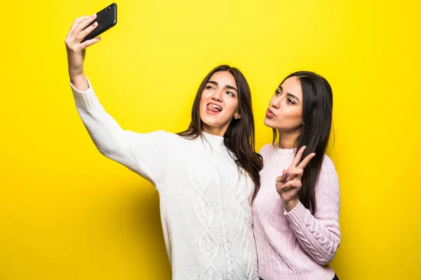 Két vidám lány portréja öltözött pulóverek, állandó, és vesz egy selfie izolált át a sárga háttér — Stock Fotó