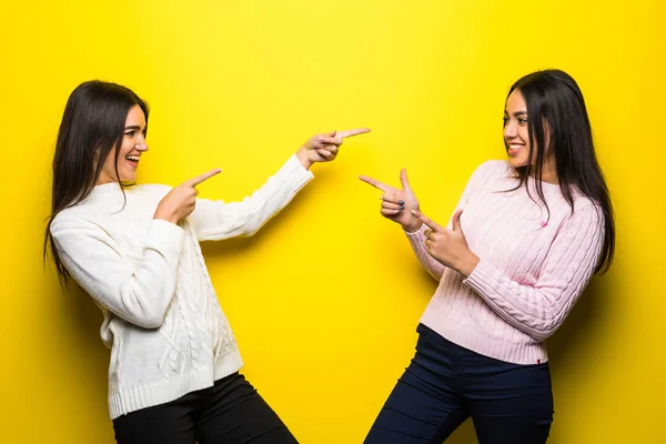 Retrato de dos chicas felices vestidas con suéteres apuntando con los dedos aislados sobre fondo amarillo —  Fotos de Stock