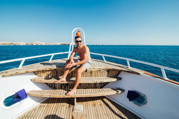 Молодой красивый мужчина в солнечных очках сидит на краю яхты и смотрит на море — стоковое фото
