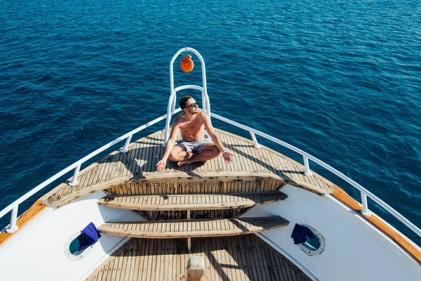 Young handsome man in sunglasses sitting at edge of yacht looking at sea — Stock Photo, Image