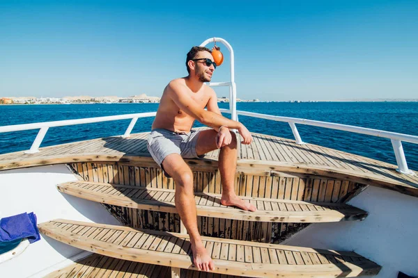 Молодой красивый мужчина в солнечных очках сидит на краю яхты и смотрит на море — стоковое фото