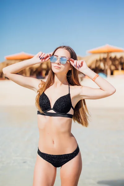 Elegant beach woman in bikini in black beachwear, sunglasses enjoying sun and sea on tropical destination during summer vacation — Stock Photo, Image