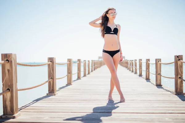 Cute, pretty young woman in a black, beautiful swimsuit walking on pierce on a sea background. — Stock Photo, Image