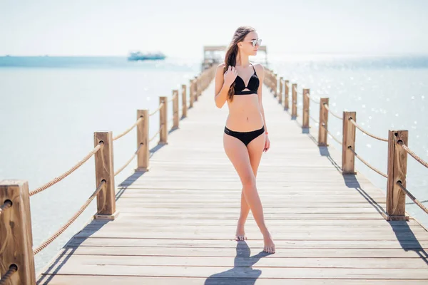 Carino, bella giovane donna in un nero, bel costume da bagno che cammina su piercing su uno sfondo marino . — Foto Stock