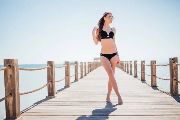 Carino, bella giovane donna in un nero, bel costume da bagno che cammina su piercing su uno sfondo marino . — Foto Stock