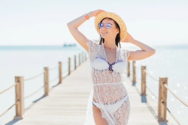 A fiatal szépség mosoly nő fehér bikini, a kalap és napszemüveg possing a háttérben tenger-kikötő — Stock Fotó