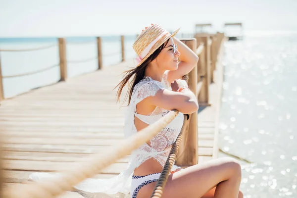 Hermosa mujer en bikini sombrero de paja y gafas de sol en el muelle de madera junto a un agua de mar —  Fotos de Stock