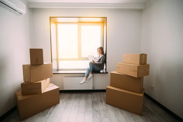 Attraktiv ung kvinna flytta, sitter på fönsterkarm bland kartonger, använder en bärbar dator och leende — Stockfoto