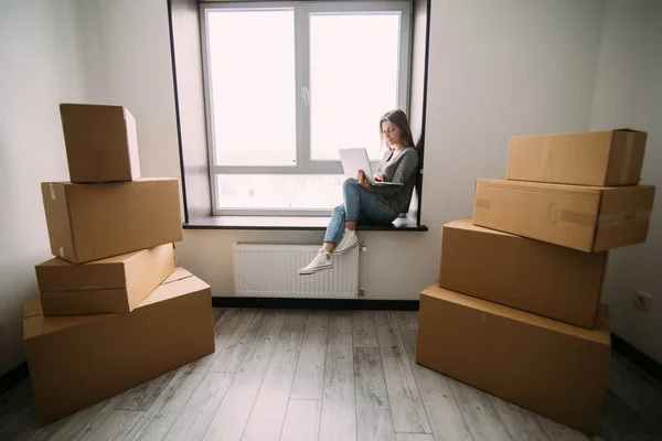 Attraktiv ung kvinna flytta, sitter på fönsterkarm bland kartonger, använder en bärbar dator och leende — Stockfoto
