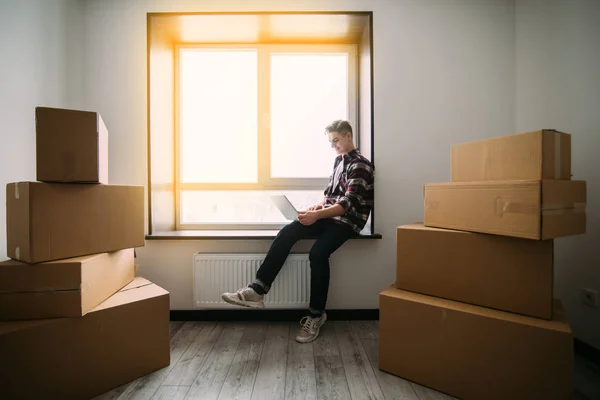 Csak költözött. Csinos, fiatal férfi ül az ablakpárkányon, és dolgozik a laptop közelében karton dobozok — Stock Fotó