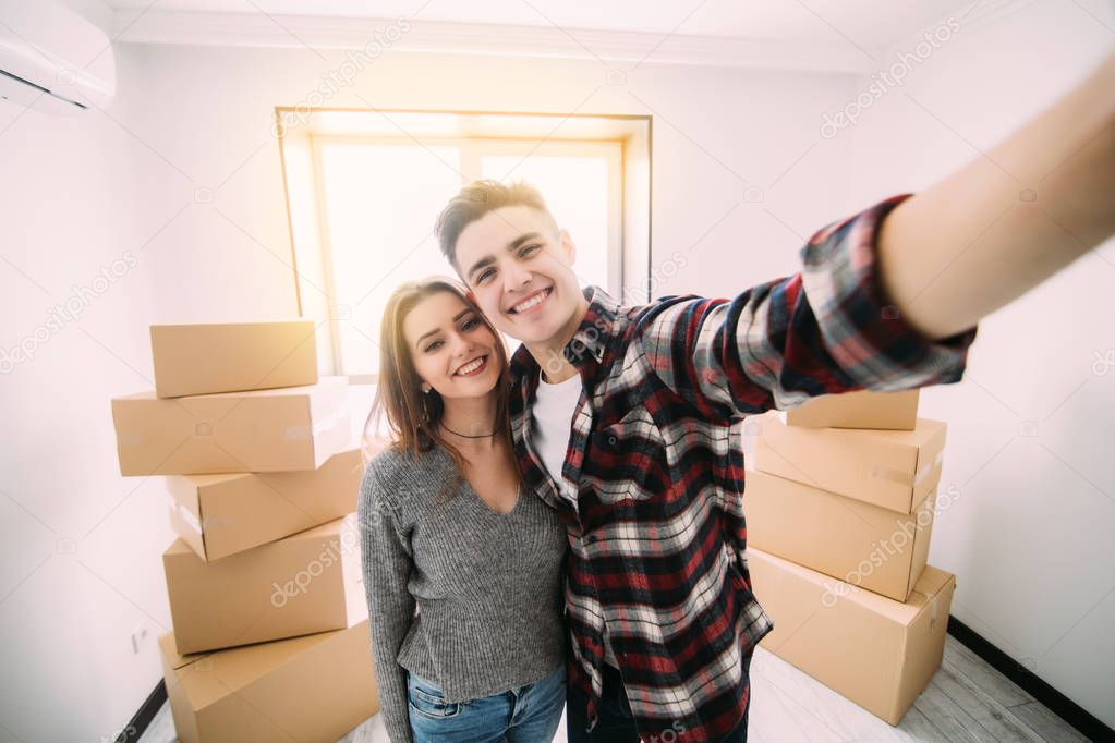Happy young couple moving to new apartment taking selfie around boxes