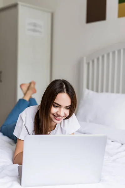 Uśmiechnięta kobieta z jej nogi lekko podniesiona przed jej laptop leżąc na łóżku. — Zdjęcie stockowe