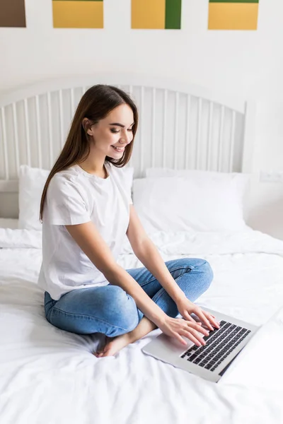 Feliz mujer hermosa casual trabajando en un ordenador portátil sentado en la cama en la casa. — Foto de Stock