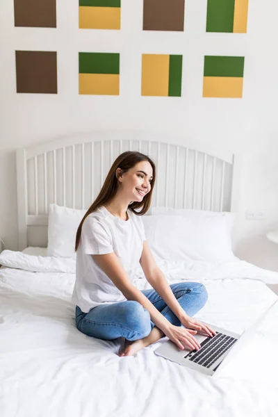 Mujer usando computadora portátil acostada en la cama. Primer plano retrato de feliz hermosa chica escribiendo en el teclado portátil, trabajando en línea en casa mientras sonríe cámara . — Foto de Stock
