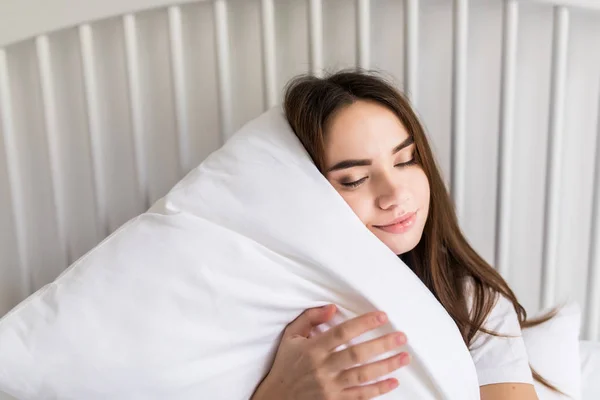 Чувственная улыбающаяся молодая женщина сидит на кровати и обнимает белую подушку — стоковое фото