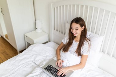 Sabahleyin yatakta otururken dizüstü bilgisayar kullanan mutlu genç esmer kadın